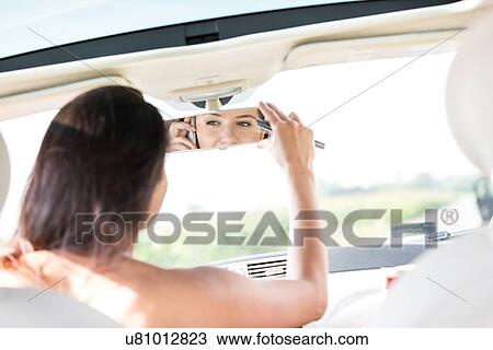 Stock Image - Woman adjusting rearview mirror while using mobile phone in car. Fotosearch