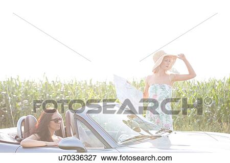 Stock Photo - Woman holding map while friend sitting in convertible against clear sky on sunny day. Fotosearch