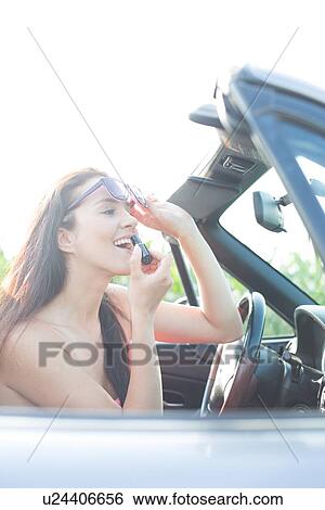 Stock Photograph - Woman holding sunglasses applying lipstick in convertible on sunny day. Fotosearch