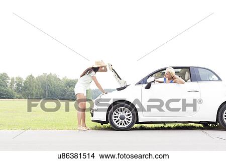 Woman looking at female friend repairing broken down car on country road View Large Photo Image Picture - Woman looking at female friend repairing broken down car on country road. Fotosearch