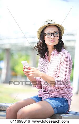 Woman looking away while holding mobile phone on bench outdoors View Large Photo Image Stock Photograph - Woman looking away while holding mobile phone on bench outdoors. Fotosearch
