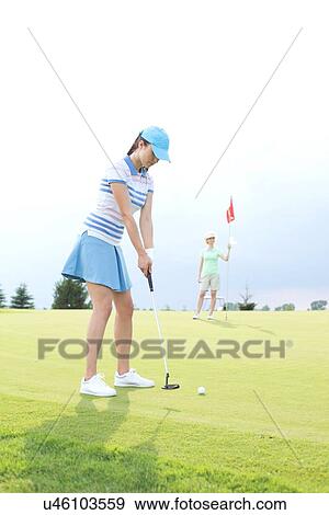 Stock Photo - Woman playing golf with female friend holding flag against sky. Fotosearch
