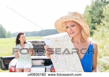 Stock Image - Woman reading map while friend leaning on convertible in background. Fotosearch