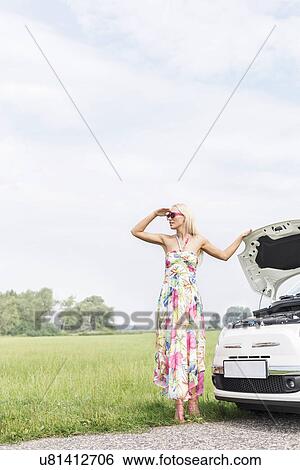 Stock Photograph - Woman shielding eyes while standing by broken down car on country road. Fotosearch
