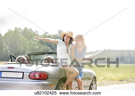 Stock Photography - Woman showing something to female friend with map while leaning on convertible. Fotosearch
