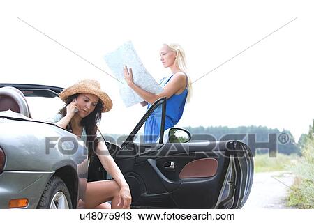 Stock Image - Woman using cell phone in convertible while friend reading map on road. Fotosearch