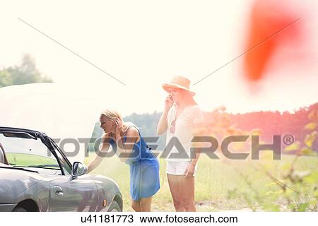 Woman using cell phone while friend examining broken down car View Large Photo Image Stock Image - Woman using cell phone while friend examining broken down car. Fotosearch