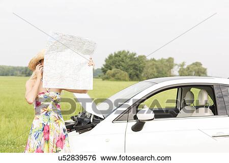 Woman using cell phone while reading map by broken down car at countryside View Large Photo Image Stock Photograph - Woman using cell phone while reading map by broken down car at countryside. Fotosearch