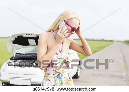 Stock Photo - Worried woman using mobile phone on country road with broken down car in background. Fotosearch