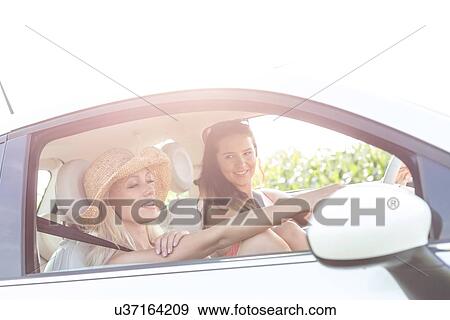 Stock Photo - Young female friends in car on sunny day against clear sky. Fotosearch