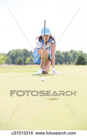 Young woman aiming ball while kneeling at golf course View Large Photo Image Stock Photograph - Young woman aiming ball while kneeling at golf course. Fotosearch