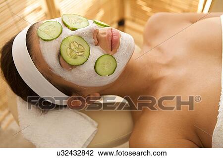 Stock Image - Young woman lying down on massage table with cucumbers on eyes and face. Fotosearch