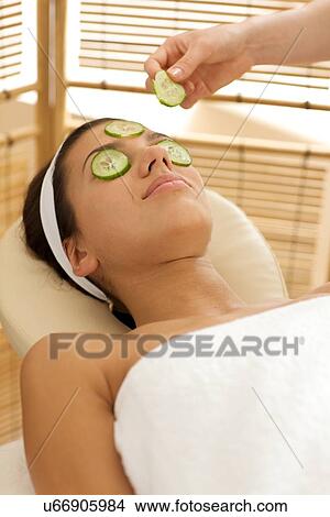 Young woman lying on massage table with cucumber slice being placed over eye View Large Photo Image Picture - Young woman lying on massage table with cucumber slice being placed over eye. Fotosearch