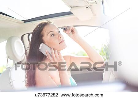 Stock Image - Young woman using mobile phone while applying mascara in car. Fotosearch