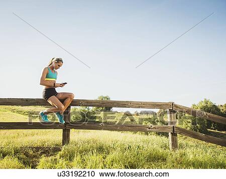 Stock Image - Woman sitting on fence, using smart phone. Fotosearch