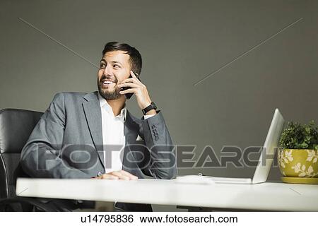 Stock Photograph - Young man sitting at desk and talking on phone. Fotosearch