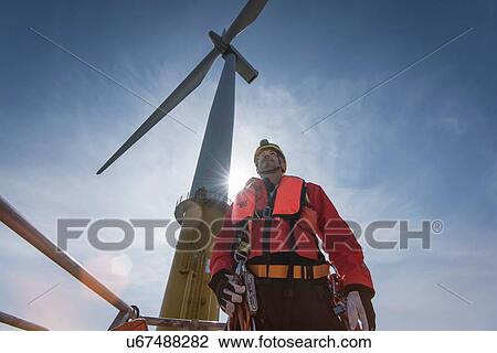 Stock Image - Engineer preparing to climb windturbine at offshore windfarm, low angle view. Fotosearch