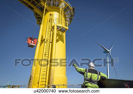 Stock Image - Engineer winches parts up to wind turbine from boat on offshore wind farm. Fotosearch