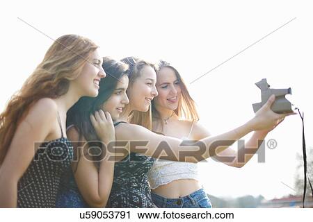 Stock Image - Four teenage girls taking instant camera selfie in park. Fotosearch