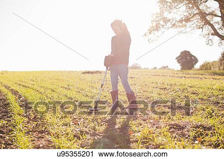 Stock Image - Girl wearing headphones searching with metal detector in sunlit field. Fotosearch