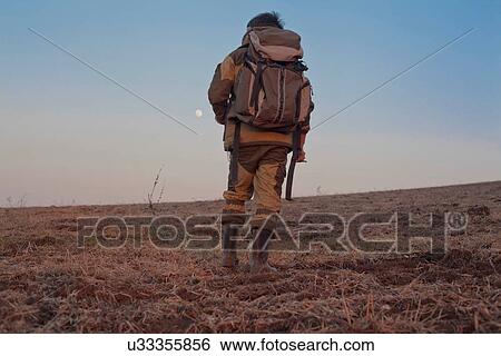 Stock Photograph - Mid adult man walking through field, rear view, low angle view. Fotosearch