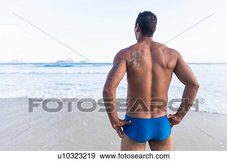 Stock Photo - Mid adult man wearing swimming trunks, looking out to sea, rear view. Fotosearch