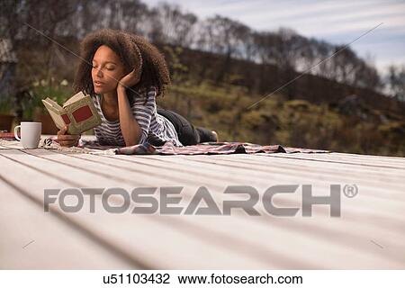 Stock Image - Mid adult woman lying down, reading book. Fotosearch