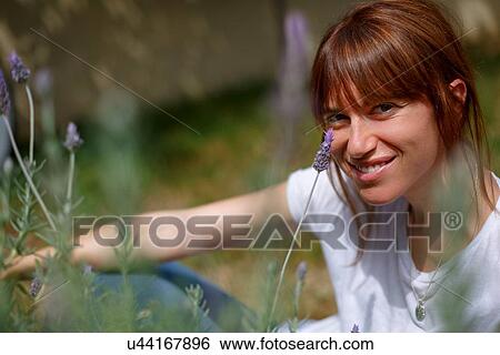 Stock Photograph - Mid adult woman sitting among lavender, looking at camera, smiling. Fotosearch