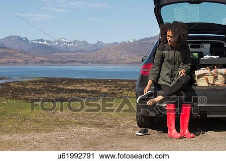 Stock Image - Mid adult woman sitting on car boot putting shoes on, Loch Eishort, Isle of Skye, Hebrides, Scotland. Fotosearch