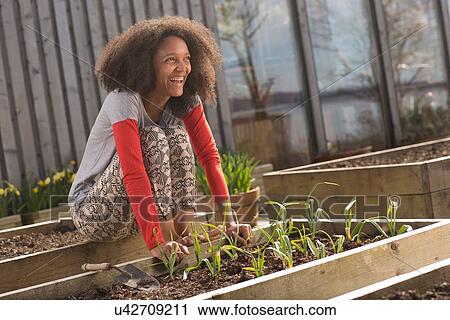 Stock Image - Mid adult woman sitting on raised bed of plants, smiling. Fotosearch