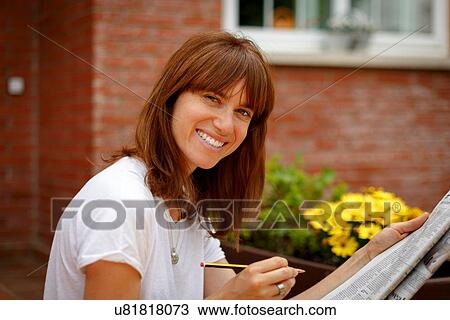 Mid adult woman sitting outdoors doing crossword, looking at camera, smiling View Large Photo Image Stock Image - Mid adult woman sitting outdoors doing crossword, looking at camera, smiling. Fotosearch