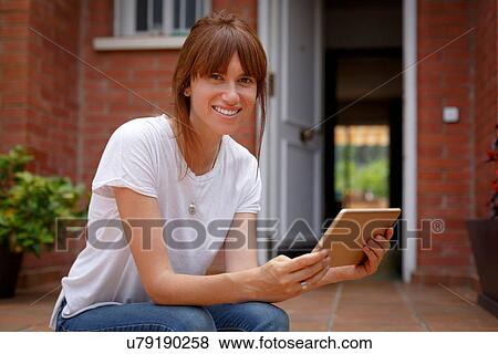 Stock Photo - Mid adult woman sitting outdoors holding digital tablet, looking at camera. Fotosearch