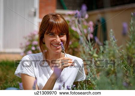 Stock Image - Mid adult woman sitting smelling lavender, smiling at camera. Fotosearch