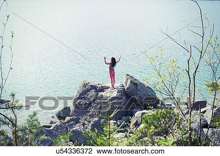 Stock Image - Mid adult woman, standing on rock beside lake, in yoga position, rear view. Fotosearch