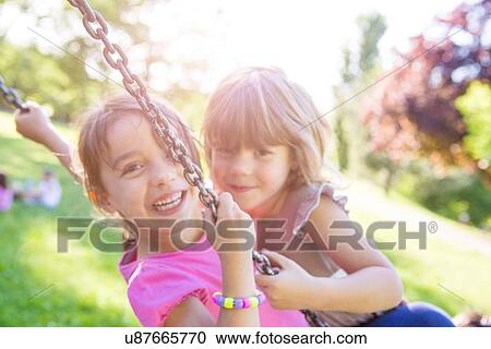 Stock Image - Portrait of two young girls swinging together on park swing. Fotosearch