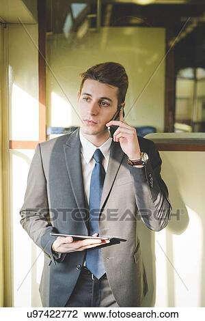 Stock Image - Portrait of young businessman commuter using digital tablet and smartphone on train.. Fotosearch
