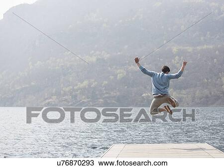 Stock Image - Rear view of young man jumping on pier, Lake Mergozzo, Verbania, Piemonte, Italy. Fotosearch