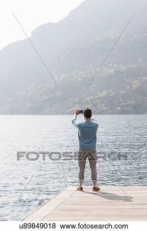 Rear view of young man taking photograph from pier, Lake Mergozzo, Verbania, Piemonte, Italy View Large Photo Image Stock Photo - Rear view of young man taking photograph from pier, Lake Mergozzo, Verbania, Piemonte, Italy. Fotosearch