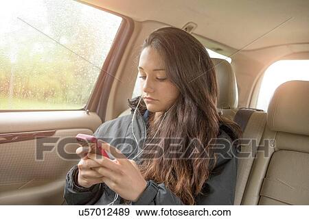 Stock Photo - Sullen teenage girl selecting smartphone music in car back seat. Fotosearch