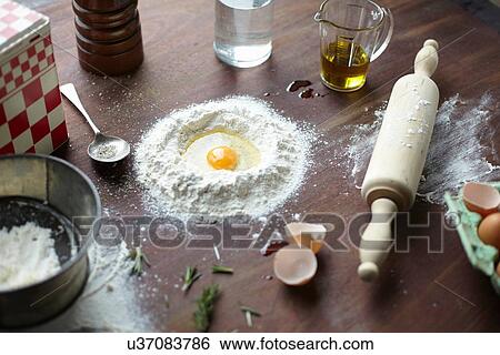 Stock Photograph - Table with raw egg in center of flour stack and kitchen utensils. Fotosearch