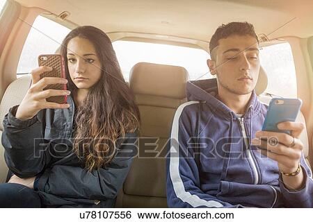 Stock Photograph - Teenage girl and young man reading separate smartphone texts in car back seat. Fotosearch