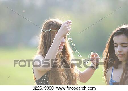 Stock Image - Two teenage girls putting on daisy chain headdresses in park. Fotosearch