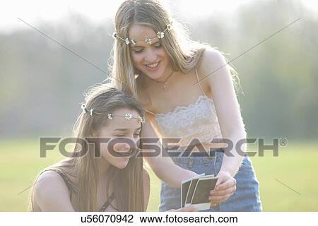 Stock Image - Two teenage girls wearing daisy chain headdresses looking at instant photographs in park. Fotosearch