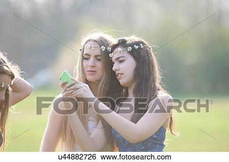 Two teenage girls wearing daisy chain headdresses reading smartphone texts in park View Large Photo Image Stock Photograph - Two teenage girls wearing daisy chain headdresses reading smartphone texts in park. Fotosearch