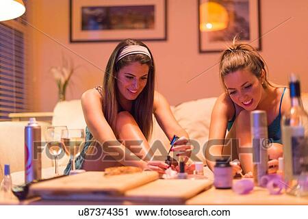 Stock Image - Two young women having girls night in, sitting on sofa, painting toes. Fotosearch