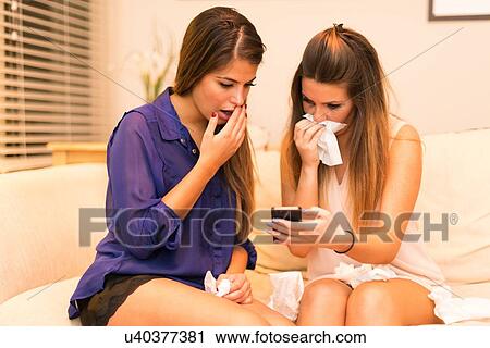 Two young women, sitting on sofa looking at smartphone, shocked expression View Large Photo Image Stock Image - Two young women, sitting on sofa looking at smartphone, shocked expression. Fotosearch