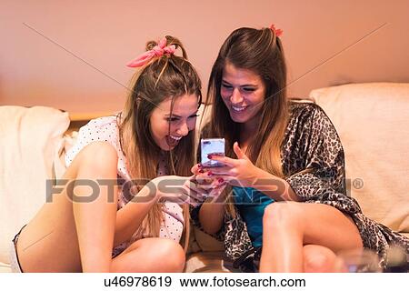 Stock Photo - Two young women, sitting on sofa looking at smartphone, laughing. Fotosearch