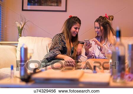 Stock Photograph - Two young women sitting on sofa, looking at smartphone, laughing. Fotosearch