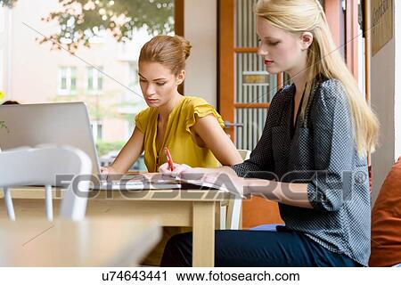 Stock Image - Two young women writing notes and using laptop in cafe. Fotosearch