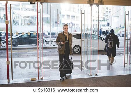 Stock Photograph - Young businessman commuter walking through glass doors with suitcase.. Fotosearch